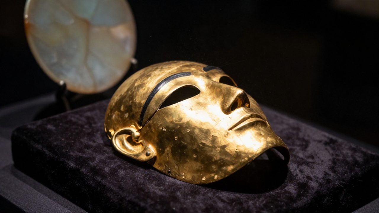 Máscara funeraria de oro antiguo con pátina sobre un cojín de terciopelo oscuro.