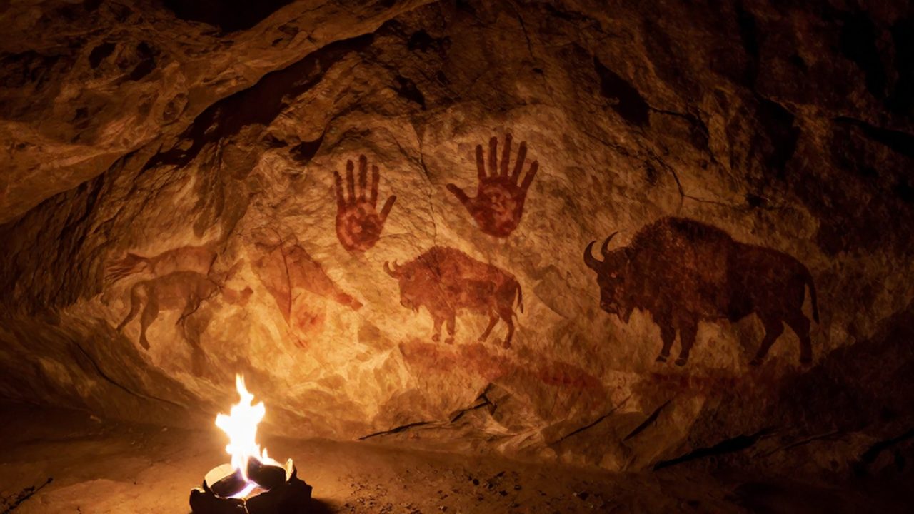 Pinturas rupestres de bisontes y huellas de manos sobre una pared de piedra iluminada por fuego.