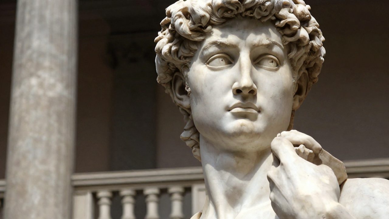 Primer plano de la escultura de mármol de David con iluminación dramática y columnas clásicas al fondo.